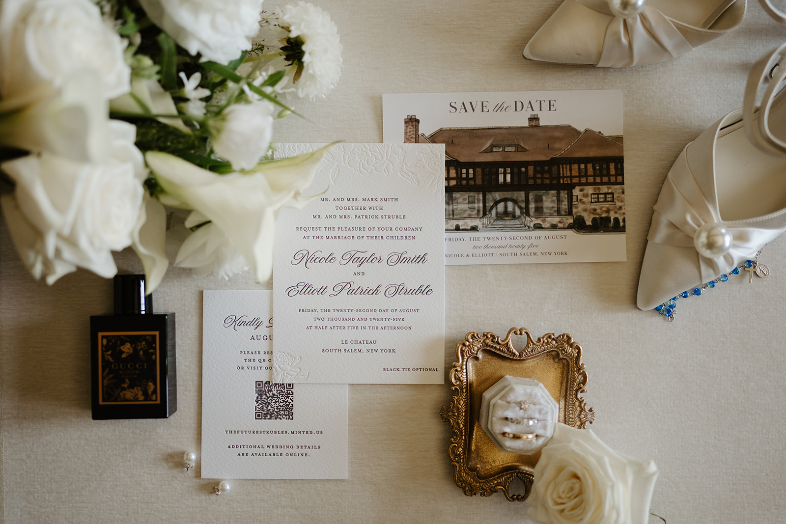 Flat lay invitation suite with silk bridal heels, rings, perfume, and white flowers.